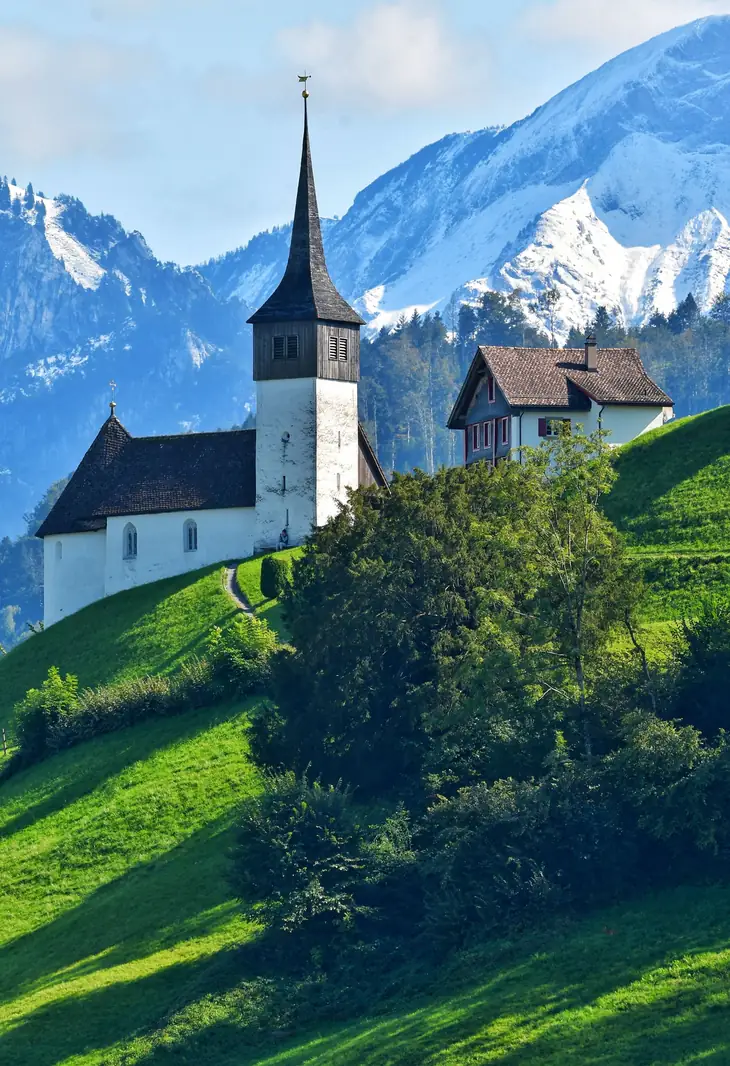 Église et bâtiments sur une croupe herbeuse face aux sommets